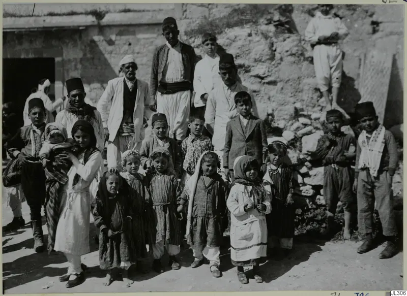 Group of people, many of them children, posed in front of a stone building and a heap of stones.