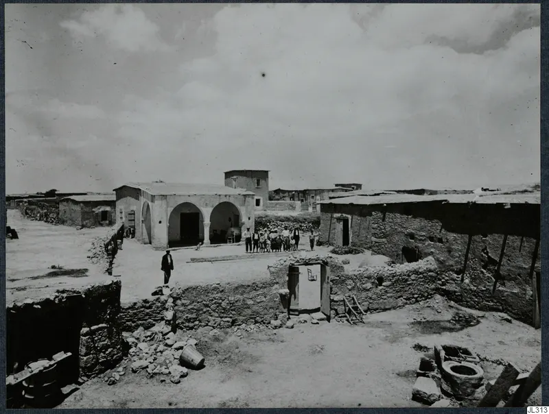 Village buildings with a whitewashed arched house, a stone wall and a group of people standing in a dusty courtyard.