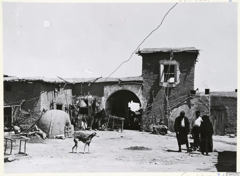 Mud-brick courtyard with a domed oven on the left, an arched gateway, a thin dog and several people