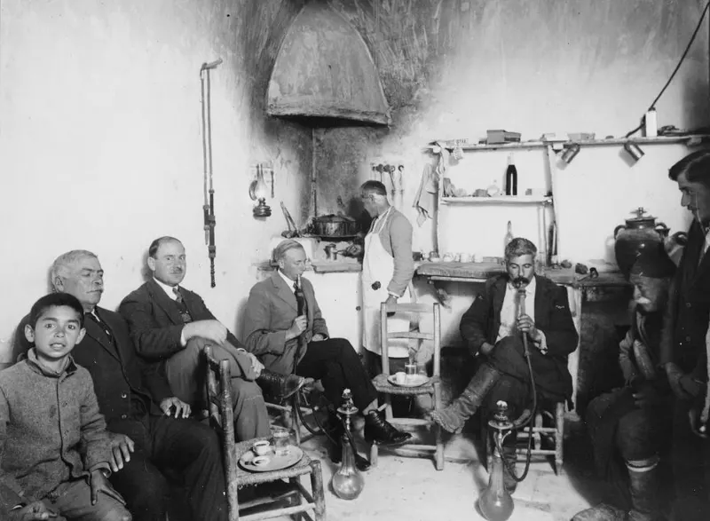 Men and a boy sit in a whitewashed café room, and a seated man at right smokes a hookah through a long hose.