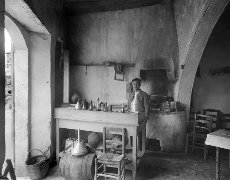Interior of a whitewashed room with a standing man, a wooden table set with metal jugs and funnels, a barrel and a basket.