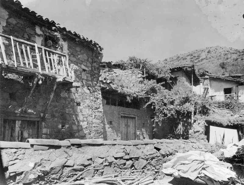 Stone houses with a wooden balcony and a low stone wall at Lazanias, with a hillside behind