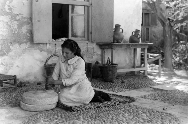 Young woman grinding grain on a hand mill in a cobbled courtyard in front of a whitewashed house wall