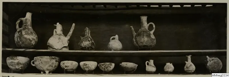 Jugs and bowls from Lapithos Markides T.9 on two shelves against a dark background.