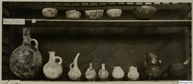 Pots and bowls from Lapithos Markides, T.15A, set out on two wooden shelves against a dark cloth.