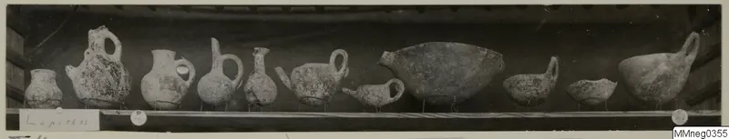Row of about a dozen jugs and bowls from Lapithos Markides, T.4 bis A, photographed on a shelf.