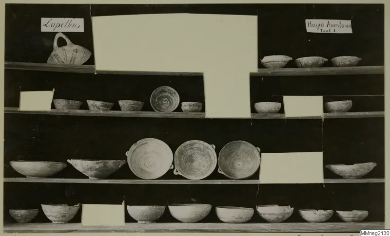 Pottery bowls and a handled jug displayed on wooden shelves, from Lapithos Markides, Tomb 1, against a dark background