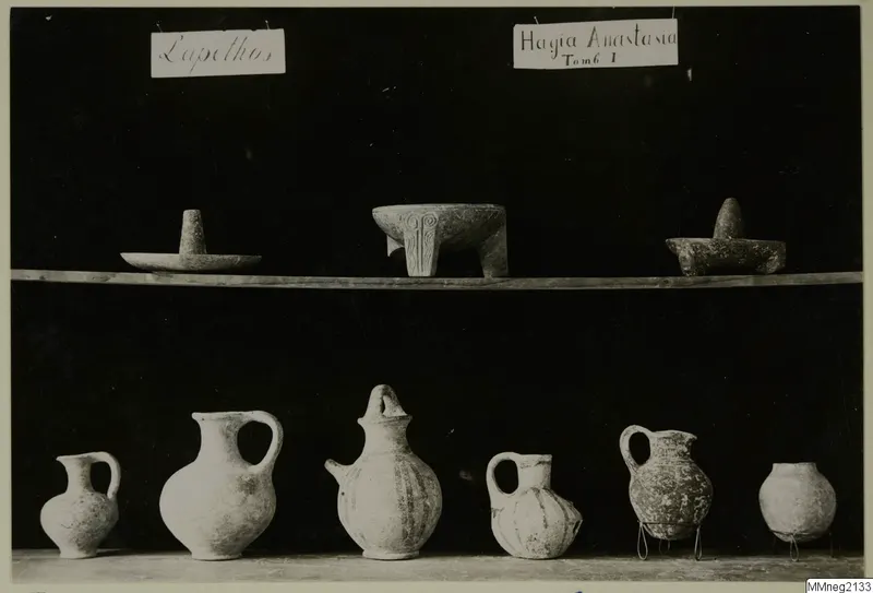 Pottery jugs and jars and small stone vessels from Lapithos, shown on two shelves against a dark background.