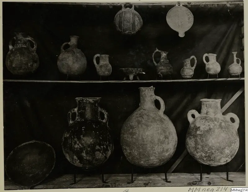 Shelved pottery vessels: small jugs and a patterned globular jar on the upper shelf, large two-handled jars on the lower shelf.