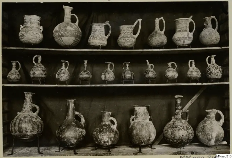Pottery jugs, juglets and jars with painted geometric patterns, arranged on three wooden shelves against a dark cloth.