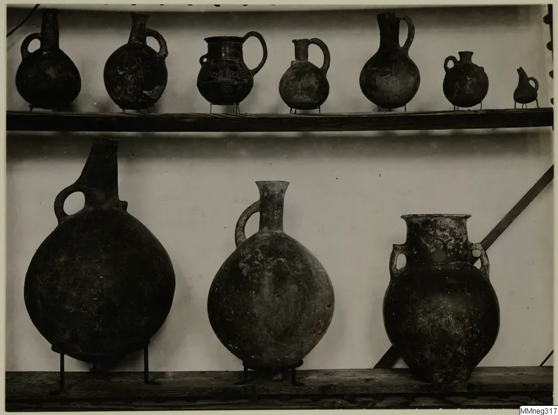 Row of small handled jugs on an upper shelf and three larger vessels on metal stands below, against a pale background