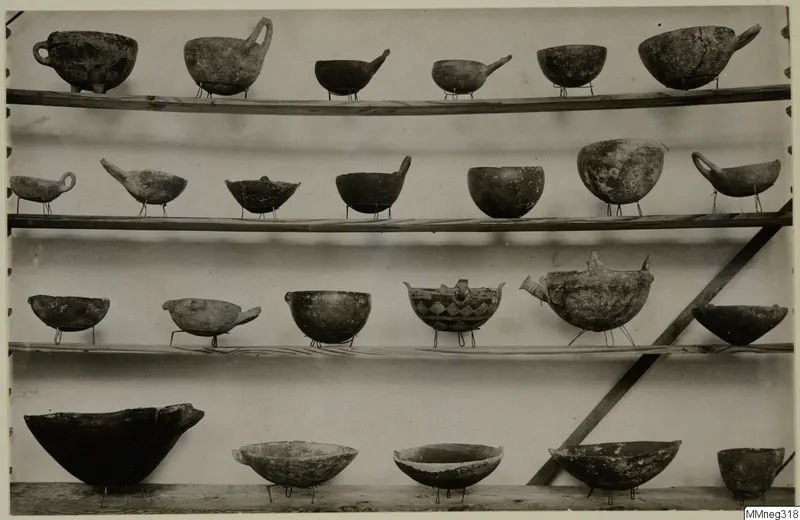 Four wooden shelves of pottery bowls and vessels with handles and spouts, some with painted patterns, each on a small wire stand.