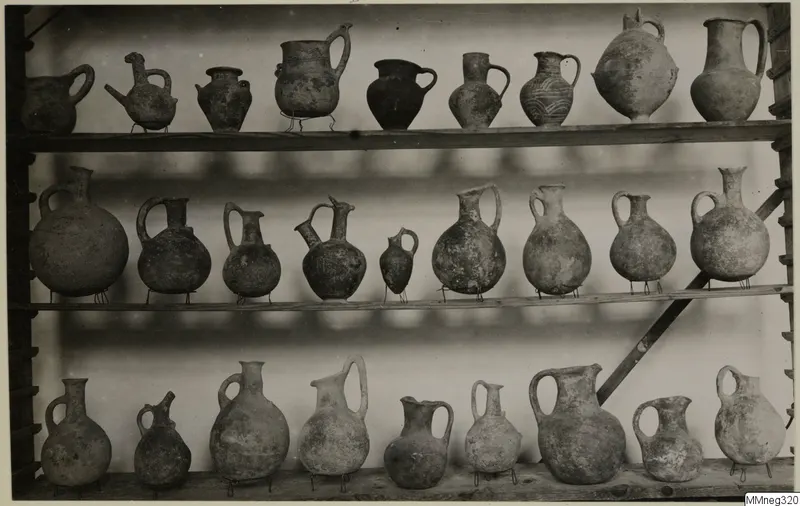 Three wooden shelves of pottery jugs and juglets, most with loop handles, a few with painted bands or patterns.