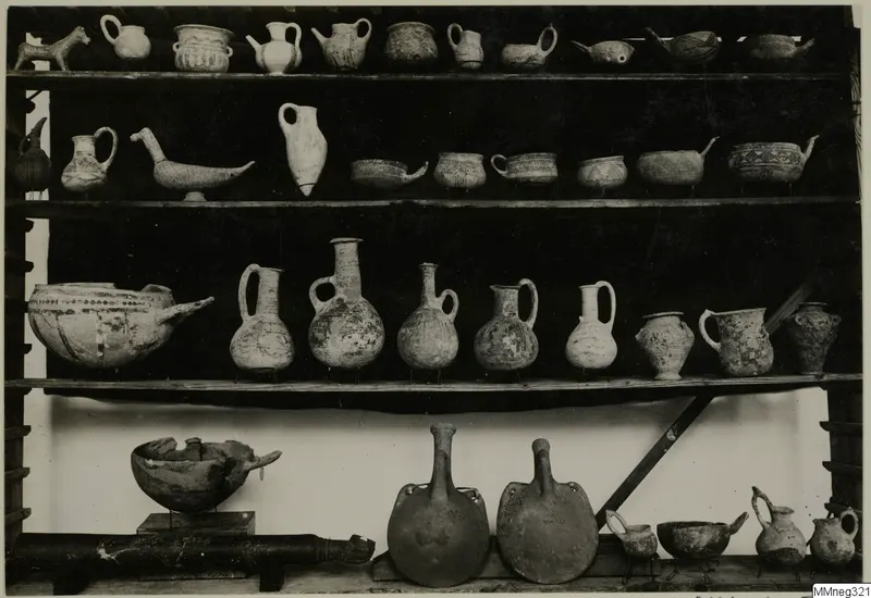 Museum shelves holding many painted pottery vessels: jugs, bowls with spouts, and two large flat-sided flasks.