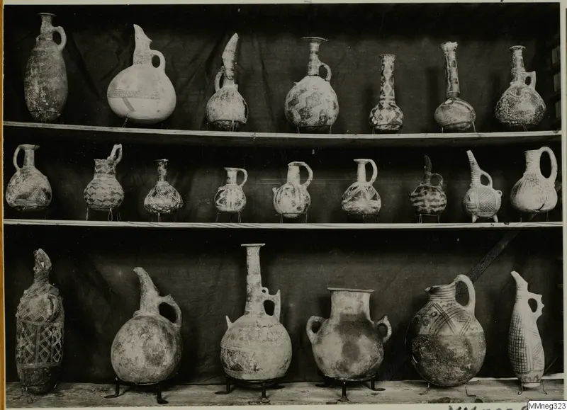 Three shelves of painted pottery jugs and juglets against a dark draped cloth