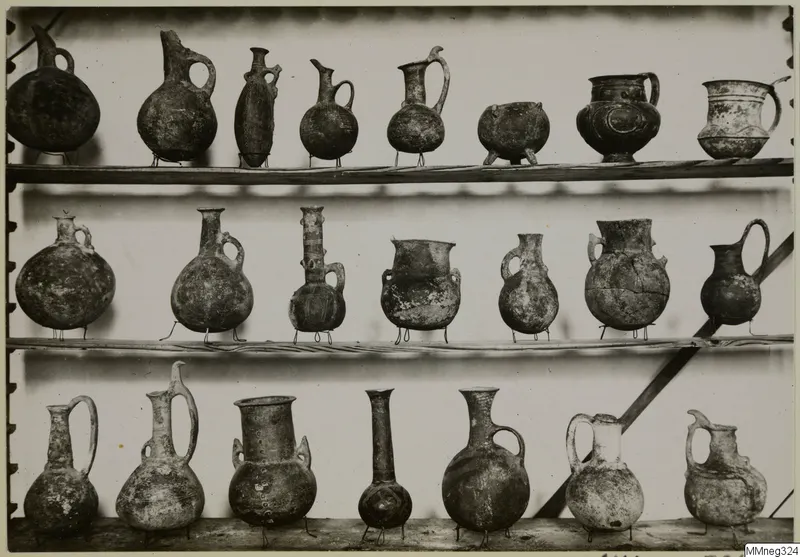 Three wooden shelves holding about twenty pottery jugs and two-handled jars, most standing on thin wire supports.