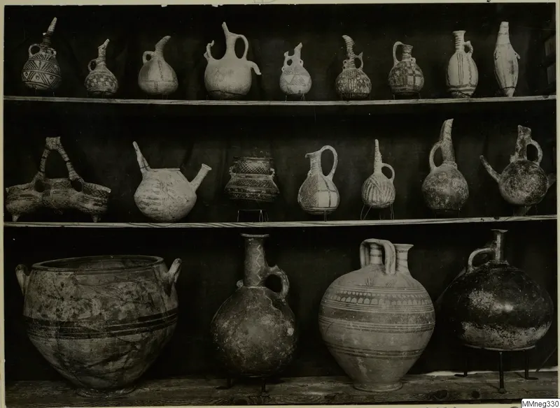 Painted pottery vessels, mostly jugs and juglets, arranged on three wooden shelves in front of a dark cloth.