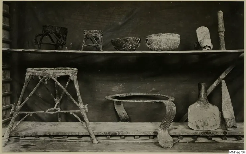 Corroded metal frames, bowls, a flask and a blade on two wooden shelves against a dark cloth.