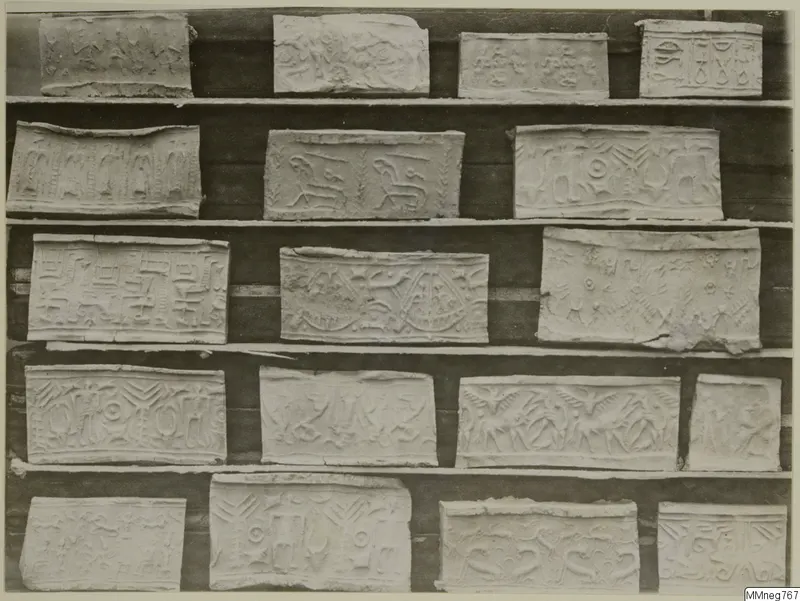 Rows of about eighteen pale rectangular pieces with shallow relief designs, set on dark shelves