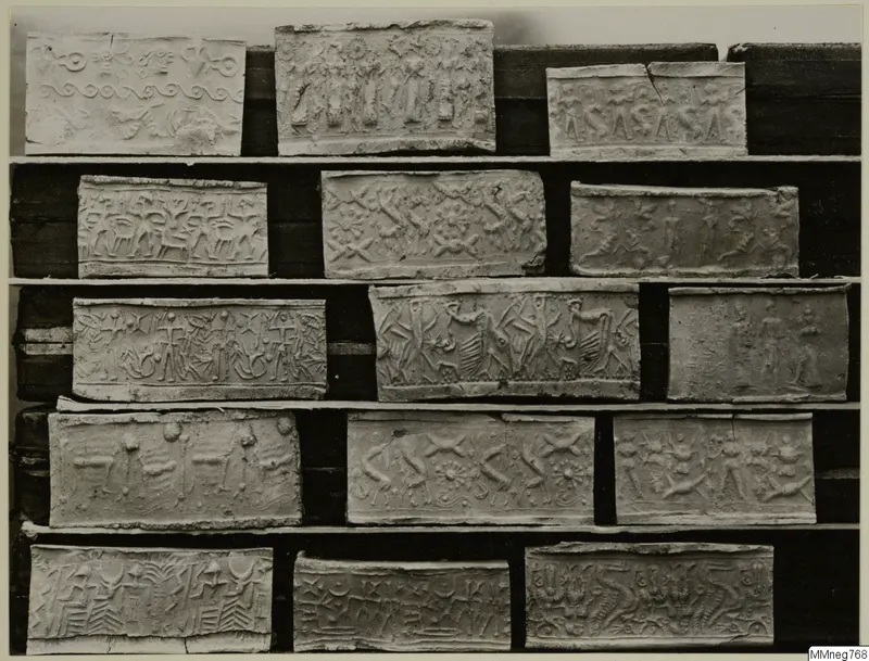 Fifteen rectangular cylinder-seal impressions with carved figures and patterns, arranged in rows on wooden shelves