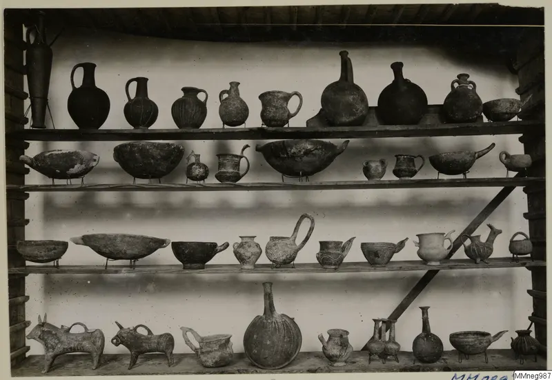 Four wooden shelves of pottery vessels on display, with jugs, flasks, bowls and two bull-shaped vessels at lower left.