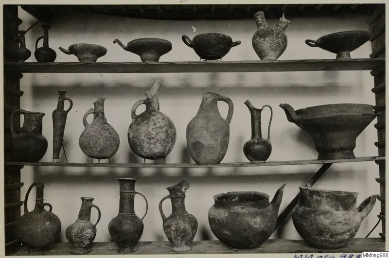 Three wooden shelves holding many pottery jugs, jars and footed bowls, displayed in the Cyprus Museum
