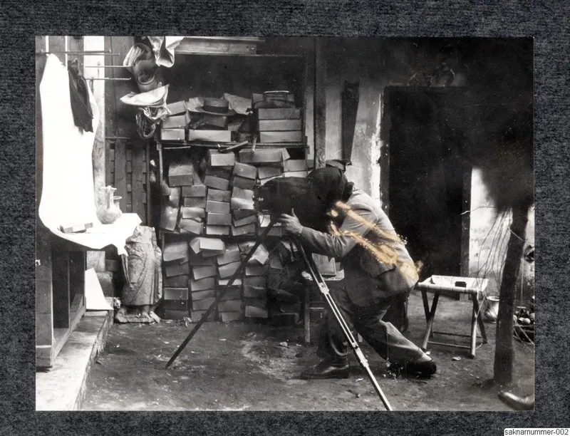 Man in a suit photographing with a large camera on a tripod, with stacked crates and boxes behind him