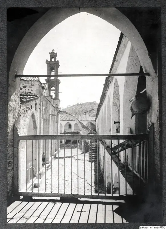 Cobbled courtyard with a church bell tower and a staircase, seen through an arched opening with iron railings