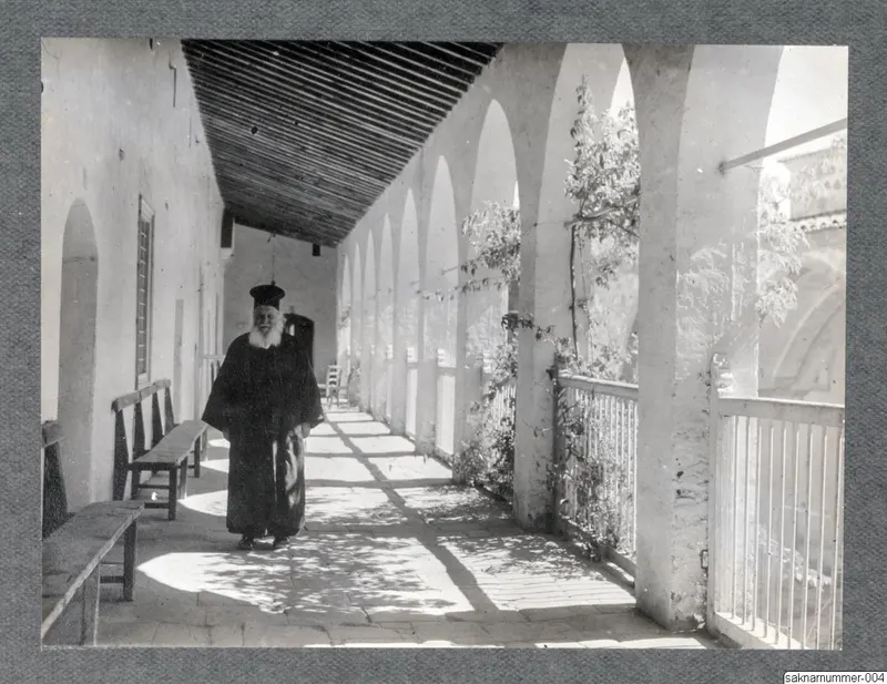 Bearded man in a long black robe and black cap standing on a paved walkway beside a row of white arches