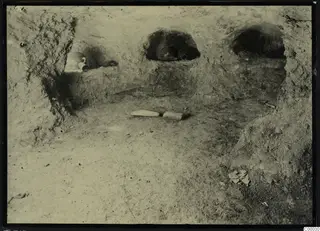 Rock-cut tomb chamber at Lapithos with vessels in wall niches and two stones on the floor during excavation