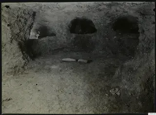 Tomb 313 a at Lapithos: interior of a rock-cut tomb chamber with arched niches, two stone slabs on the floor and a vessel in a niche