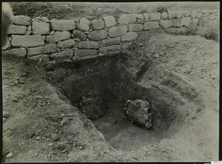 Excavated pit of Tomb 301 at Lapithos, with a dry-stone wall along its far edge and large rocks inside