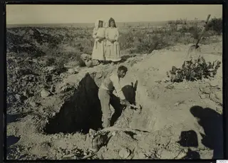 Excavation trench at Lapithos with a man digging and two standing figures, beside a shovel and a pick.