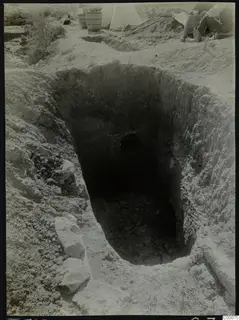 Tomb 313 at Lapithos: excavated rectangular shaft with a dark opening in the far wall, a kneeling worker and baskets beside it.
