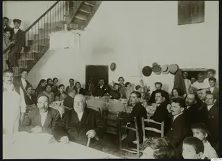 Many people of different ages seated around two long tables set with flowers and tablecloths, most looking toward the camera.