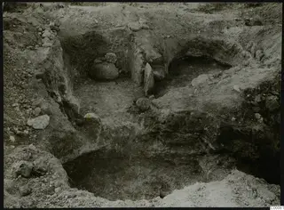 Excavation of Tombs 320 and 323 at Vrysi tou Barba, Lapithos, showing cut earth walls, stones and dry grass at the edges.