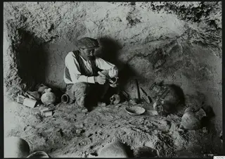 Man in a cap brushing pottery among vessels inside a rock-cut tomb chamber