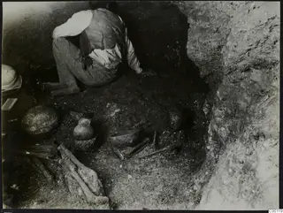 Tomb 306 c. at Lapithos: a man kneels among pottery vessels and bones in an excavated tomb chamber.