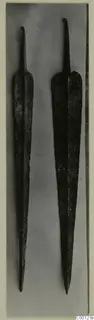 Two long metal blades with narrow tangs and pointed tips, lying side by side on a grey backdrop.