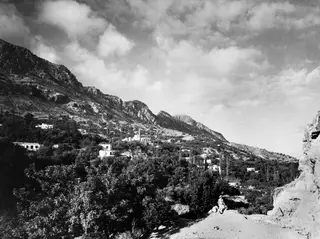 Landscape view of Lapithos, with white houses on a wooded mountain slope and a seated figure in the foreground