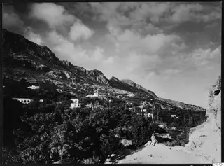 Landscape view of Lapithos, with houses on a hillside below steep rocky mountains and a single figure in the foreground