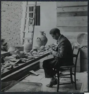 Erik Sjöqvist seated at a table covered with pottery sherds and jugs, working on the pieces, in Nicosia