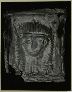 Embossed metal plaque with a raised face framed by a ridged headdress; the sheet is crumpled and torn at the edges