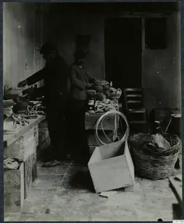 Two men in caps working among pottery vessels, crates and a wicker basket in a bare storeroom