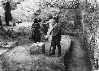 Excavation trench at Soli with workers holding picks and shovels beside a large cut stone block and a stone wall