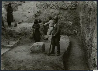 Workers with a pick and shovels in a stone-walled excavation trench, with an ancient wall rising behind them.