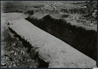 Excavation trench at Soli with a dark soil face, a pale cleaned floor, loose stones and spoil heaps behind.