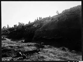 Excavation of the theatre at Soli, with people standing and sitting along the top of an earth bank and stone wall.