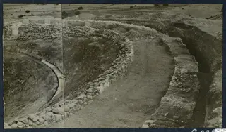 Excavated curved stone walls of the Theatre at Soli, with a trench, spoil heaps and trees beyond.