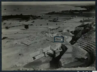 Elevated view of the excavated theatre at Soli, with curved stone seating, cut stone blocks and the sea with ships beyond.